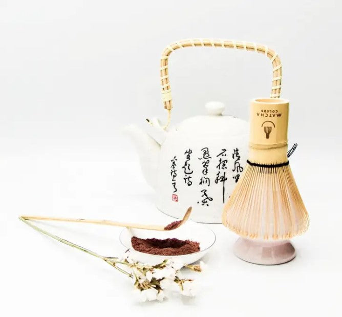 Cuchara medidora de bambú (Chashaku) de Matcha colors para servir matcha. La cuchara se encuentra sobre un bol con polvo de matcha cacao/chocolate, junto a una tetera y un batidor chasen de Matcha Colors.