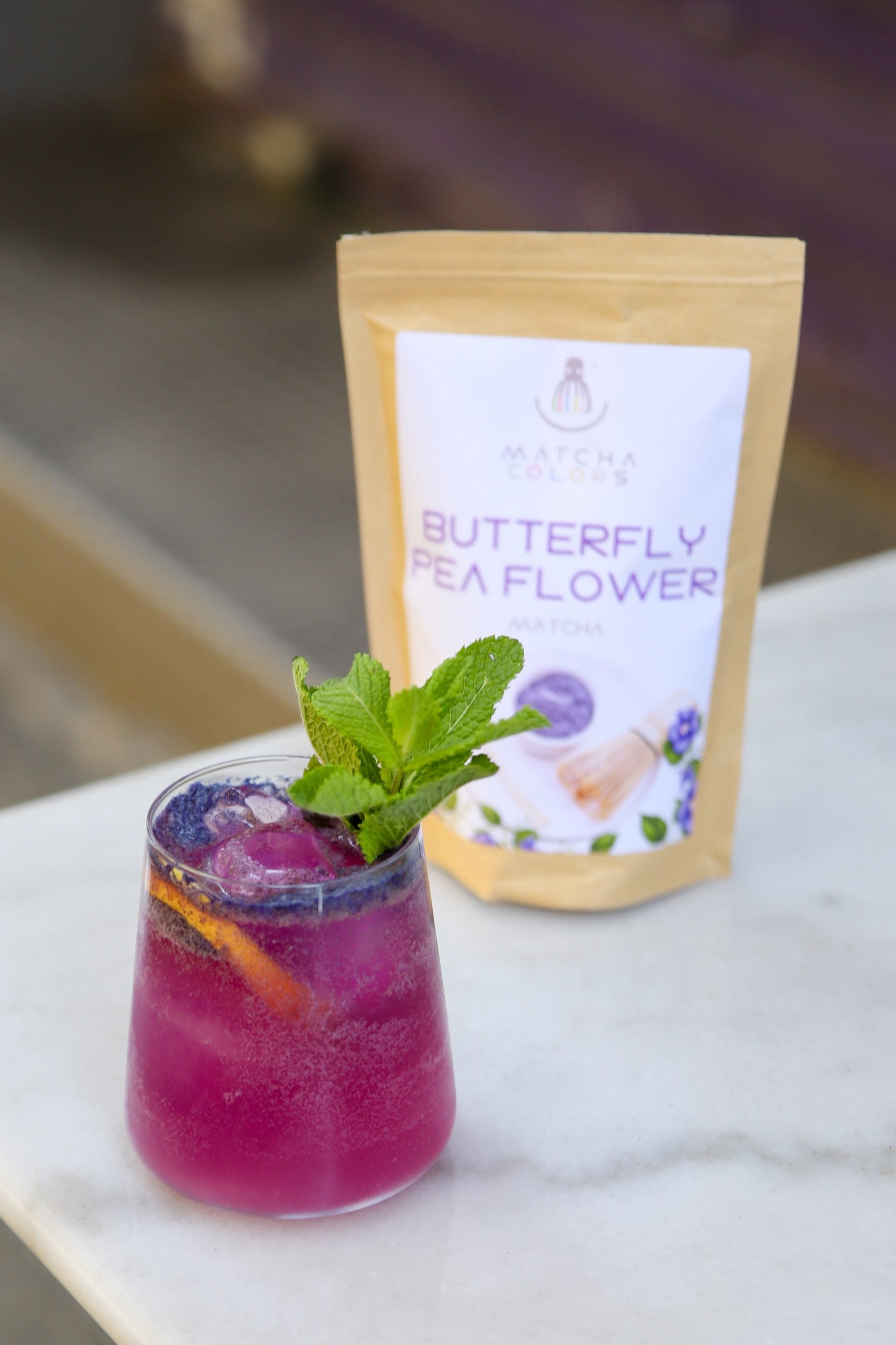 A vibrant purple cocktail garnished with mint, shown next to the Matcha Colors Butterfly Pea Flower powder pouch.